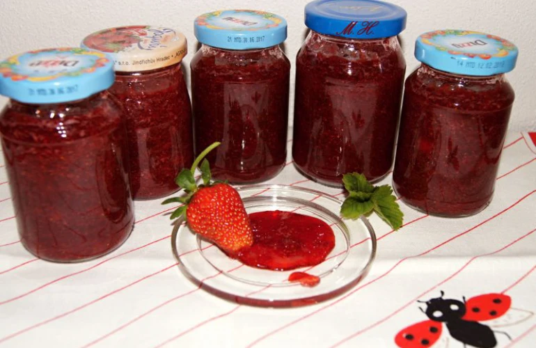 Sklenice domácí jahodové marmelády s čerstvou jahodou na talíři.