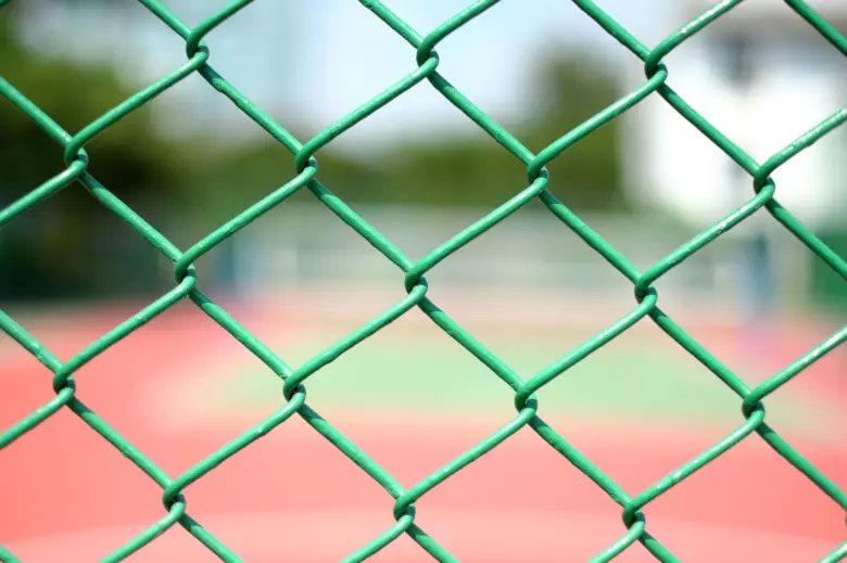 Zelený drátěný plot s pletivem před sportovním hřištěm.