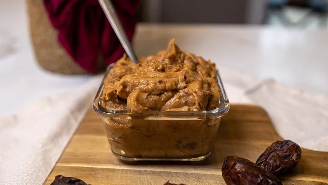 Datlová pasta v skleněné misce s lžící a daty vedle na dřevěném prknu.
