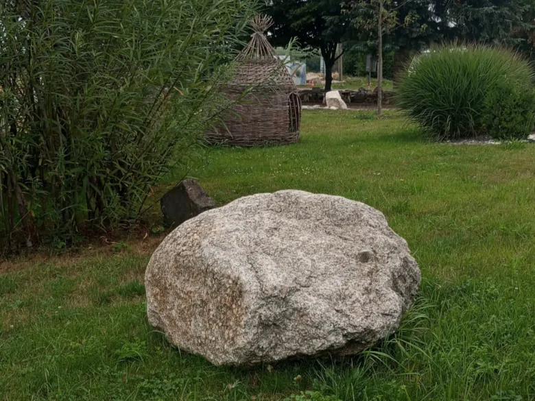 Velký přírodní kámen v zahradě mezi trávou a keři.