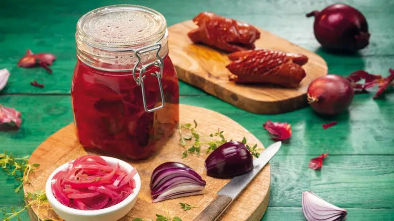 Nakládaná červená cibule v sklenici s čerstvě nakrájenou cibulí na dřevěném prkénku.
