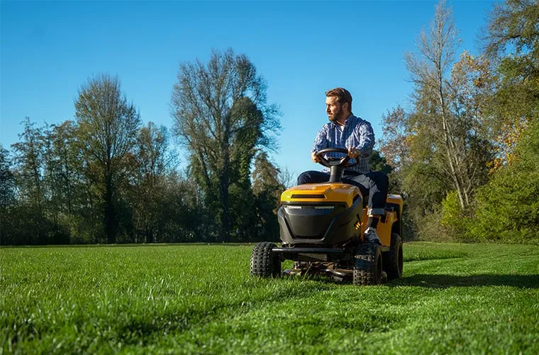 Muž řídí žlutý traktor na trávníku v zahradě.