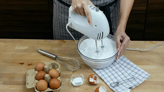 Osoba šlehá bílky vajíček elektrickým mixérem na stolu.