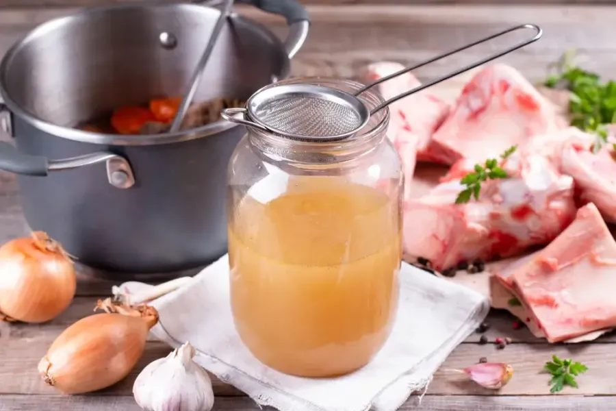 Domácí kostní vývar v sklenici s surovými ingrediencemi na dřevěném stole.