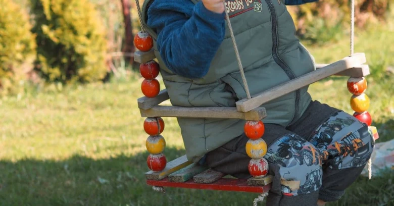 Dítě sedí na dřevěném houpačce v zahradě s barevnými koulkami.