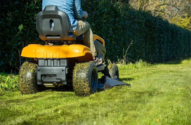 Žlutý zahradní traktor s řidičem kosí trávu v záhonu.
